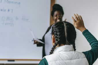 Smiling African American female teacher standing near whiteboard and looking at schoolgirl raising hand